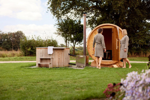 Two people in bathrobes enjoying a barrel sauna and outdoor hot tub in the green yard of a lodge.