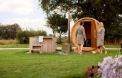 Two people in bathrobes enjoying a barrel sauna and outdoor hot tub in the green yard of a lodge.