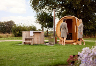Dos personas con albornoces disfrutan de una sauna de barril y jacuzzi exterior en el jardín de una cabaña.