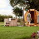 Two people in bathrobes enjoying a barrel sauna and outdoor hot tub in the green yard of a lodge.