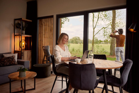Mujer trabaja en una laptop en una cabaña moderna mientras un hombre observa la naturaleza por la ventana.
