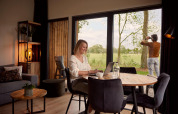 Kvinde arbejder på bærbar computer ved bord i moderne lodge, mens mand kigger ud i naturen gennem vindue.