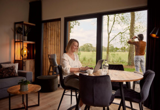 Mujer trabaja en una laptop en una cabaña moderna mientras un hombre observa la naturaleza por la ventana.