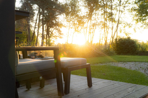 Coucher de soleil à un lodge avec terrasse en bois, fauteuils extérieurs, table et vue sur le jardin arboré.