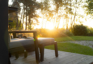 Atardecer en un lodge con terraza de madera, conjunto de asientos, mesa y vista a jardín y árboles verdes.