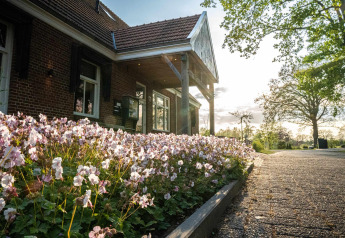 Foto van een lodge met een voortuin vol roze bloemen, bestrate oprit en bomen bij zonsondergang.