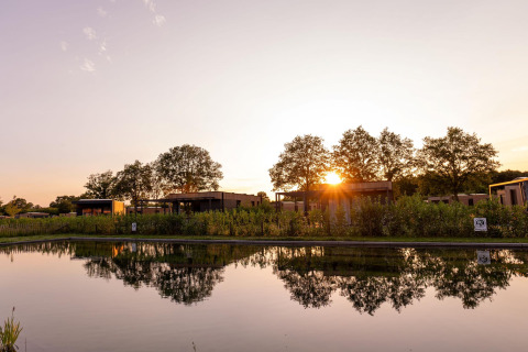 Solnedgang over en lodge ved en sø, moderne hytter og træer spejler sig i vandet, fredelig natur.