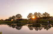Sunset over a lodge by a pond, modern cabins and trees reflecting in the water, peaceful natural setting.