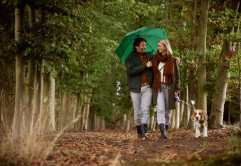 Par går tur med hund under grøn paraply i skov ved en lodge, smilende og iført efterårstøj.