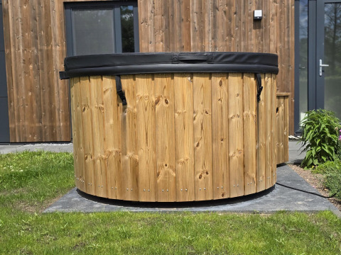 Bañera de hidromasaje de madera al aire libre con tapa negra junto a una cabaña moderna en los Países Bajos.