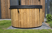 Bañera de hidromasaje de madera al aire libre con tapa negra junto a una cabaña moderna en los Países Bajos.