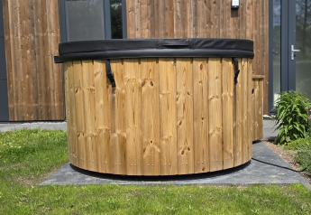 Bañera de hidromasaje de madera al aire libre con tapa negra junto a una cabaña moderna en los Países Bajos.