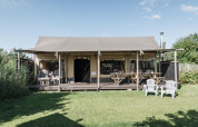 A photo of a cozy lodge with a shaded porch, picnic tables, plastic chairs, and a green lawn outside.