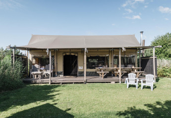A photo of a cozy lodge with a shaded porch, picnic tables, plastic chairs, and a green lawn outside.