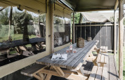 Udendørs spiseområde ved en lodge med træbord, bænke, vinflaske og to glas i landlige omgivelser.