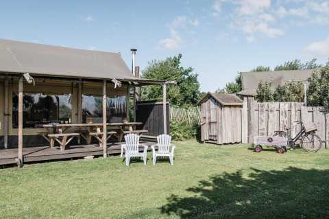 Escena exterior de una cabaña con terraza de madera, bancos de picnic, dos sillas y césped verde.