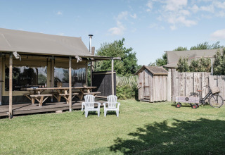 Escena exterior de una cabaña con terraza de madera, bancos de picnic, dos sillas y césped verde.
