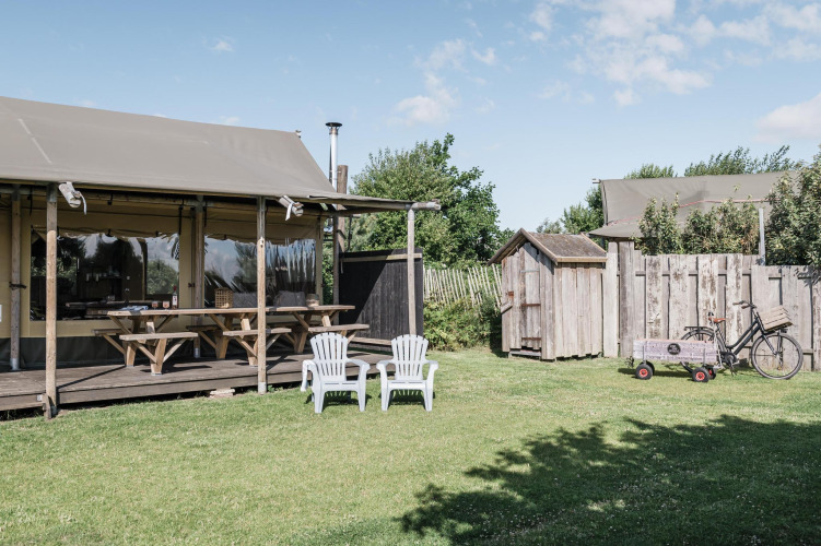 Outdoor lodge scene with a wooden deck, picnic benches, two white chairs, a bike, shed, and green lawn.