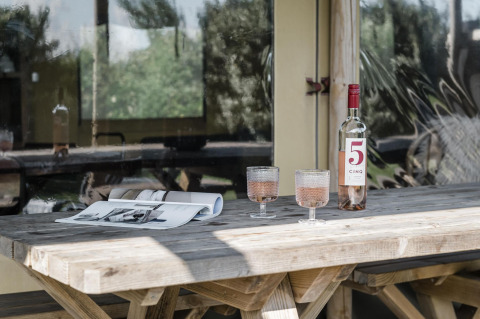 Mesa rústica de madera con una botella de vino rosado, dos copas y una revista abierta junto a una cabaña.