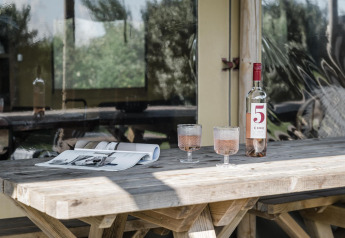 Mesa rústica de madera con una botella de vino rosado, dos copas y una revista abierta junto a una cabaña.