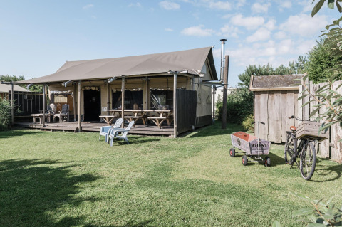 Helles Lodge mit überdachter Terrasse, Holzbänken, Stühlen, Fahrrad und Karren im grünen Garten.