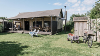 Lodge lumineux avec terrasse couverte, tables de pique-nique, chaises, vélo et chariot sur pelouse verte.