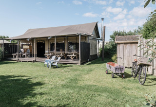 Helles Lodge mit überdachter Terrasse, Holzbänken, Stühlen, Fahrrad und Karren im grünen Garten.