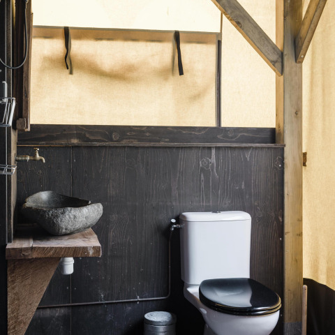 Baño rústico en una cabaña con lavabo de piedra, estructura de madera y aseo, rodeado de paredes de lona.