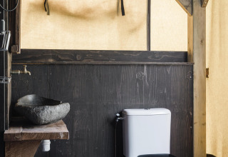 Et rustikt badeværelse på en lodge med stensvask, trækant og toilet, omgivet af trævægge og lærred.