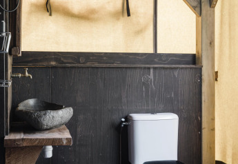 Salle de bain rustique dans un lodge avec lavabo en pierre, structures en bois et toilettes, entourée de toile.