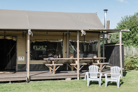 Buitenfoto van een lodge met overdekt terras, twee picknicktafels en twee witte stoelen op het gras.