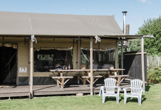 Udendørs billede af et lodge med veranda, to picnicborde og to hvide plaststole på græsplæne foran.