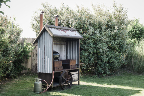 Casa de chapa ondulada en un jardín con chimenea, ruedas y garrafa de gas al aire libre entre arbustos.