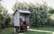 A corrugated iron outdoor shed with chimney, wheels, and gas bottle, standing in a lush green garden.