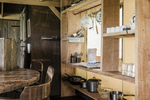 Cocina rústica de una cabaña con estanterías de madera, ollas, utensilios, tazas y una mesa de madera.