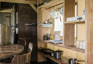 Cocina rústica de una cabaña con estanterías de madera, ollas, utensilios, tazas y una mesa de madera.