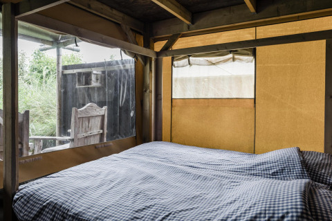 Innenansicht einer gemütlichen Lodge mit Doppelbett, karierten Bettbezügen und Blick ins Grüne.