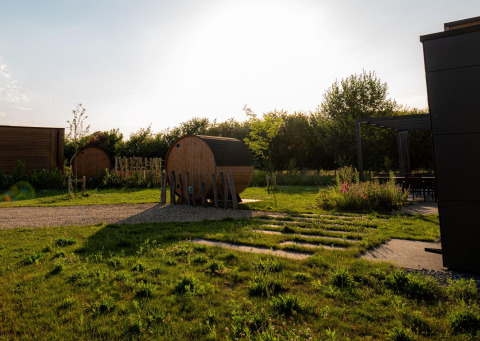 Außenbereich einer Lodge mit Fasssaunas, grüner Wiese, Terrasse und Sonnenuntergang.