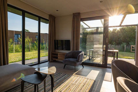 Salón moderno de lodge con grandes ventanales, luz solar y vistas al jardín y terraza exterior.