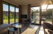 Salón moderno de lodge con grandes ventanales, luz solar y vistas al jardín y terraza exterior.