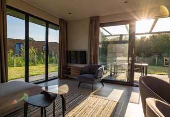 Salón moderno de lodge con grandes ventanales, luz solar y vistas al jardín y terraza exterior.