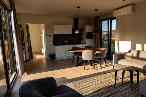 Sollys strømmer ind i en moderne lodge med stue, køkken, spisebord og store vinduer ud til naturen.