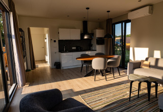 Sollys strømmer ind i en moderne lodge med stue, køkken, spisebord og store vinduer ud til naturen.
