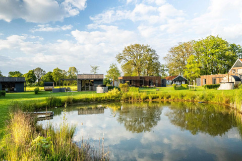Lodges aan een vijver met groene grasvelden en bomen, onder een deels bewolkte blauwe lucht.