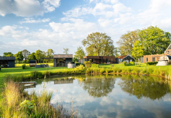 Lodge moderne près d'un étang, entouré de pelouse et d'arbres, sous un ciel bleu partiellement nuageux.