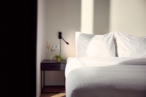 Et solrigt hotelværelse med en hvid seng, to puder og et natbord med planter og en væglampe.
