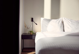 Et solrigt hotelværelse med en hvid seng, to puder og et natbord med planter og en væglampe.