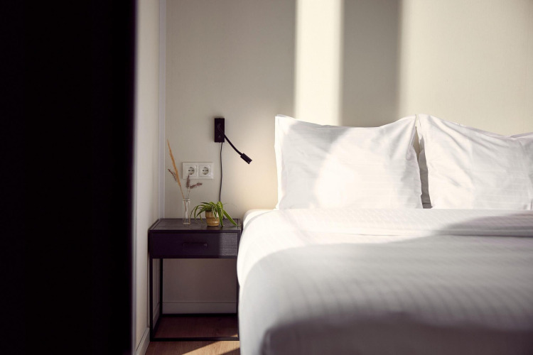 Habitación luminosa de una posada con cama blanca, dos almohadas y una mesita con plantas y lámpara.