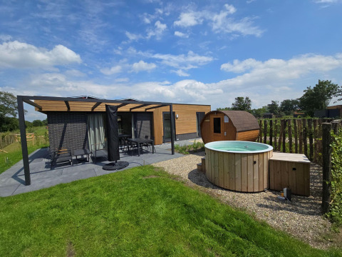 Vista exterior de una cabaña moderna con terraza, muebles de jardín, sauna de barril y jacuzzi redondo.