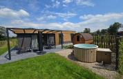 Vista exterior de una cabaña moderna con terraza, muebles de jardín, sauna de barril y jacuzzi redondo.